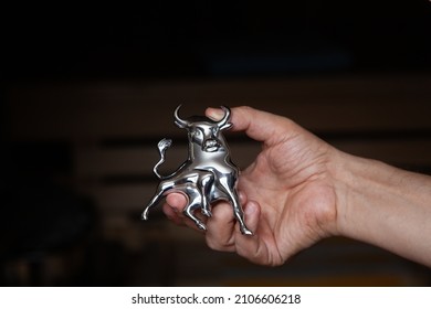 Right Hand Holds A Chrome Bull Figurine With Horns And A Raised Tail On A Black Background

