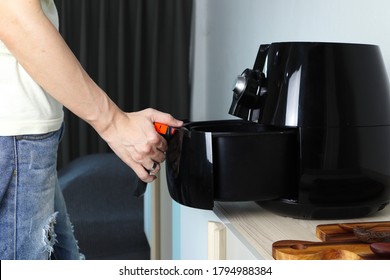 Right hand of man holding the tray of the black deep air fryer or oil free fryer appliance which is on the wooden table in the white kitchen ( air fryer ) during lunch with family members