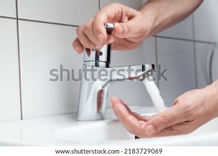 The right hand opens the tap on the lever faucet above the white tiled sink. A jet of water pours on the left hand. White tiles on the wall



