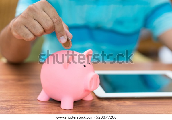 Right hand putting\
coin into piggy saving bank with tablet computer background, copy\
space on Right side.