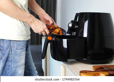 Right hand putting the fried chicken into the tray of the black deep fryer or oil free fryer appliance which is on the wooden table in the white kitchen ( air fryer )