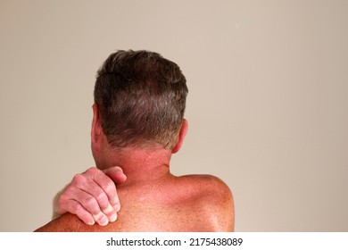 Right hand of a shirtless adult white man massaging his left shoulder as seen from a rear view. Close-up of a caucasian guy rubbing his left shoulder with his right hand in front of a wall indoors