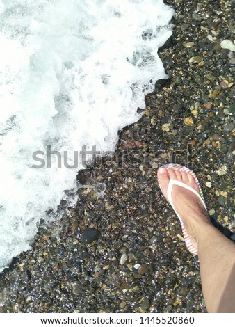 the right leg of a man in a white rubber slipper for swimming stands on the mottled surface of sea pebbles at the coastal edge with white foam of the incoming wave, diagonal co-position, space for tex [[stock_photo]] © 