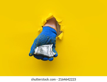A right man's hand in a blue fabric work glove holds crumpled dollar bills (money). Torn hole in yellow paper. Concept for gastarbeiter, handyman, salary for job. Copy space.