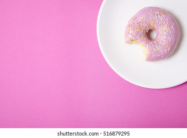 A ring donut with pastel frosting and sprinkles on a bright pink background, with a bite missing and blank space at side