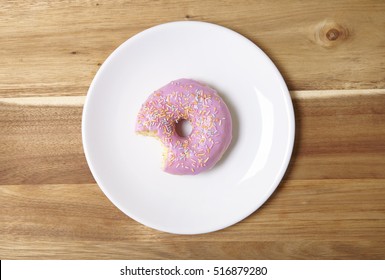 A ring donut with pastel pink frosting and sprinkles on a wooden kitchen counter background, with a bite missing