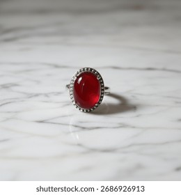 
​Silver ring featuring a large, oval, deep red cabochon stone, possibly garnet or ruby, set on a white marble surface.