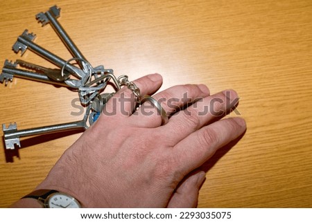 Ring on ring finger and a bunch of keys chained to it. The keychain lies on a wooden table next to the hand. Unfree Man, Duties of Marriage