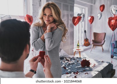 Ring is perfect! Attractive young woman looking at engagement ring with smile while her boyfriend proposing her in the bedroom