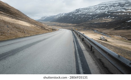 The Ring Road sweeps through a broad North Iceland valley, snow-dusted peaks rising on both sides above an abandoned turf farmhouse in soft spring light.