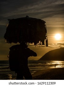 Rio De Janeiro, Rio De Janeiro, Brazil - 12 13 2019: A Hawker Carries His Products Overhead Using An Umbrella, As He Walks Along The Beach In Late Afternoon