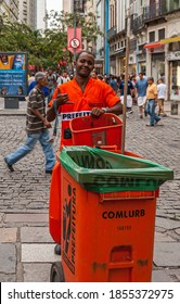 Rio De Janeiro, Brazil - December 26, 2008: El Centro District. Colored City Garbage Collector In Orange Garb Shows The Shaka Sign With His Right Hand In Front Of His Rolling Trash Can. Street Scene 