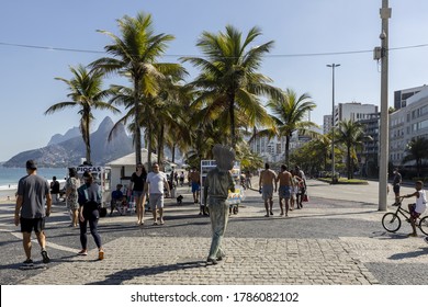 RIO DE JANEIRO, BRAZIL - Jul 19, 2020: People Strolling Around In Face Masks Tom Jobim Bronze Statue Passing By Street Vendors And Palm Trees On Ipanema Beach