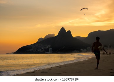 Rio De Janeiro, Brazil - July 2nd 2016: Ipanema Beach With Silhouette Of Young Brazilian Boy Kicking A Football & Paraglider Overhead At Sunset & Dois Irmãos (Two Brothers) Mountains In Background