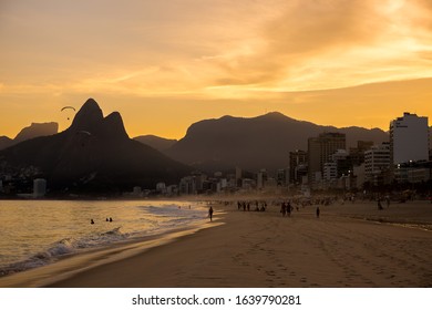 Rio De Janeiro, Brazil - July 2nd 2016: Ipanema Beach With Silhouette Of People Relaxing & Having Fun & Paragliders Overhead At Sunset & Dois Irmãos (Two Brothers) Mountains In Background