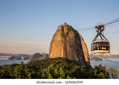 Rio De Janeiro, Brazil - July 4th 2016: Sugarloaf Mountain In Guanabara Bay, Urca At Sunset With Warm Sky Overhead & Tourists In Cable Car Heading Down From The Top While Enjoying The Views In Brazil