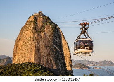 Rio De Janeiro, Brazil - July 4th 2016: Sugarloaf Mountain In Guanabara Bay, Urca At Sunset With A Warm Sky Overhead And Tourists In A Cable Car Heading To The Top While Enjoying The Views In Brazil