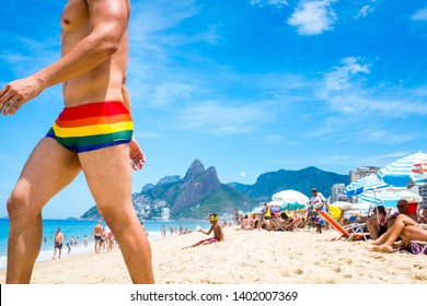 RIO DE JANEIRO - CIRCA JANUARY, 2017: Gay man in rainbow pride swimsuit walks past beachgoers relaxing on a bright summer day in Ipanema Beach.