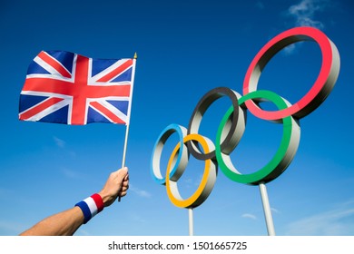 RIO DE JANEIRO - MARCH, 2016: A Hand Of A Team GB Supporter Wearing Red White And Blue Sweatband Holds A UK Flag In Front Of Olympic Rings.