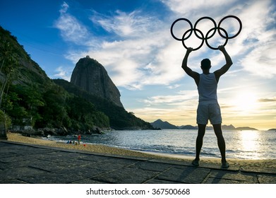 RIO DE JANEIRO - OCTOBER 31, 2015: Silhouette Of Athlete Holds Olympic Rings In Front Of A Sunrise Of Sugarloaf Mountain At Red Beach, Praia Vermelha In Urca. 