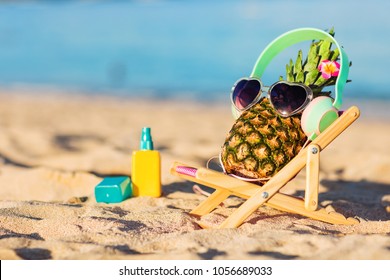 Ripe attractive girl-pineapple in stylish headphones lying on sunbed on the sand against turquoise sea. Listening music, relaxing. Wearing sunglasses. Tropical summer vacation concept. Sunbathing
