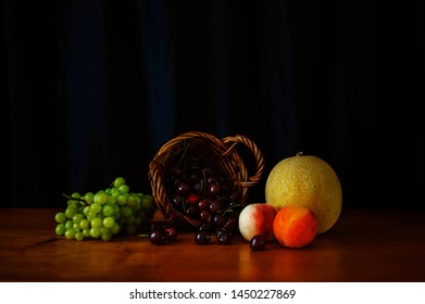 Ripe, Red Cherries In A Woven Basket, A Juicy Bunch Of Green/white Grapes, A Yellow Honeydew Melon And Bright Orange Peaches. Shot On A Farmhouse Table Top With Natural Light. Classic Still Life Style