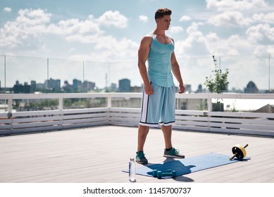 Ripped Male Is Standing On Roof Of High Building In Downtown. He Is Getting Ready For Training With ABS Roller And Dumbbells. Guy Is Having Bottle Of Water For Drinking During Work Out