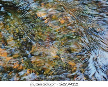 Ripples in the river flow through the rocks, colorful in the summer in bright sunlight, have background of the shiny pebbles, at the bottom of river In a abstract, form of pebbles below water surface.