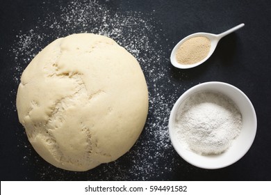 Risen Or Proved Yeast Dough For Bread Or Pizza On Floured Slate Surface, Ingredients On The Side, Photographed Overhead With Natural Light (Selective Focus On The Of The Dough And The Flour In Bowl) 