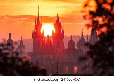 The rising sun in between towers of the Church of Our Lady before Tyn in UNESCO site Prague seen through leaves.