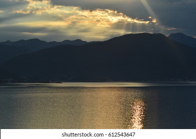 The Rising Sun Is Breaking Through Clouds Above A Mountain And Onto The Sea. A Small Village Can Be Seen At The Water's Edge.