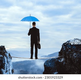 Risks and challenges of entrepreneurship. Businessman with portfolio and umbrella standing on rope over abyss in mountains, back view