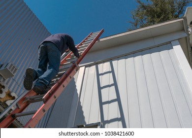 Risks Of A Worker Climbing Uninsured Ladder To Work On The Roof Of A House On A Beautiful Sunny Day With Blue Sky.