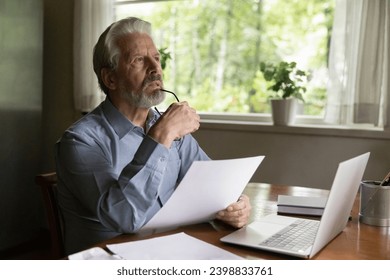 Risky business. Thoughtful aged man ponder on document at office table before laptop screen. Pensive elderly male sit at workplace hold paper letter look away planning deal result analyze information - Powered by Shutterstock