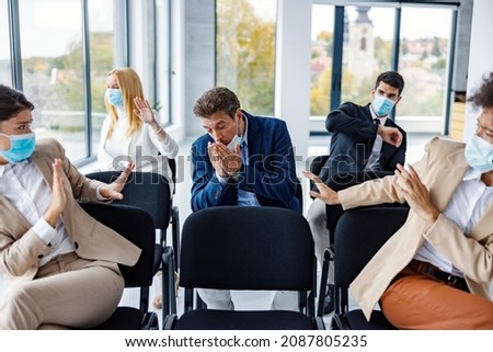 Risky symptoms on seminar between business people. A businessman sneezing and coughing in the boardroom while his colleagues with face masks defending themselves from a deadly virus.