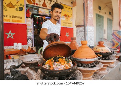 Rissani, Morocco - September 18th, 2019: Young moroccan man cooking tajine with vegetables in the street, and showing it to the tourist to sell meal, in Rissani, Morocco.