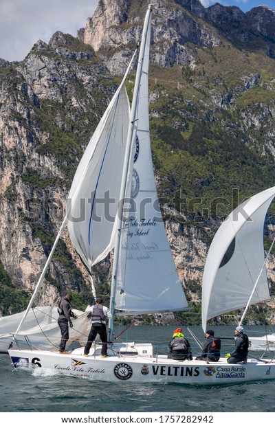 Riva-del-Garda, Italy - 28042019: scenic\
mountain landscape sailing competition, heeling sailing boat in\
motion on foreground, sailing crew team on deck, green hills of\
Riva-del-Garda on\
background