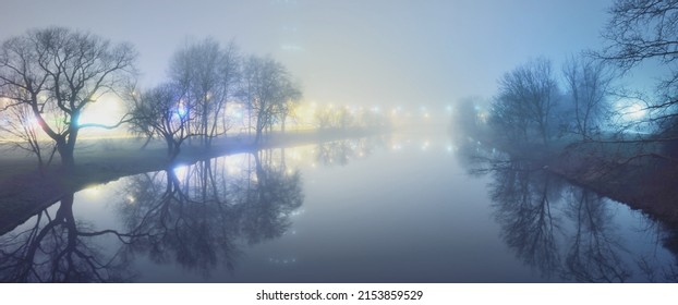 River and a city park in a fog at night. Bright illumination by the street lanterns. Symmetry reflections on the water. Riga, Latvia. Dark cityscape
