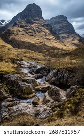 The River Coe flows through Glen Coe with The Three Sisters mountains in early spring, Scotland