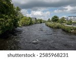 The river Corrib flowing through Galway, Ireland, with modern buildings on one bank and a pathway with lush greenery on the other, under a partly cloudy sky