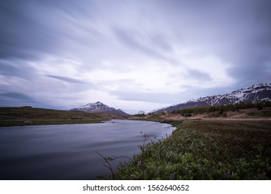 A River Flowing Out Of Eyjafjörður Fjord In Northern Iceland. Mountainous Landscape And Flowing Clouds In The Distance