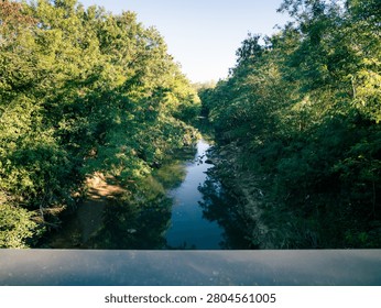 River flows between tall green trees. Sunlight filters through dense foliage. View from a bridge overlooks the waterway. Calm, natural landscape ideal for travel themes