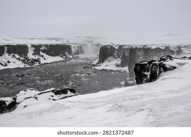 A river flows forcefully between dark basalt cliffs covered with snow. In the background, the majestic Selfoss waterfall is partially frozen, immersed in a desolate and cold arctic landscape during a winter snowfall under a white sky.