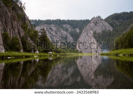 A river flows gently between massive rocky cliffs, one side covered in lush green moss, the other side rugged and grey. The sky is overcast as the calm water gently laps against the shore.