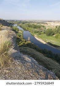 A river flows through green fields and trees under a clear sky. The landscape shows hills and a diverse terrain, indicating a rural setting. The sunlight brightens the scene.