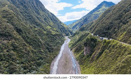 A river flows through a steep valley surrounded by green hills. A road runs along the edge of the valley. The scene is bright and clear under a blue sky with clouds.