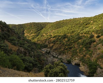 A river flows through a valley surrounded by green hills. The sun shines on the landscape. Some trees and rocks are visible along the riverbank. The sky shows clouds and lines.