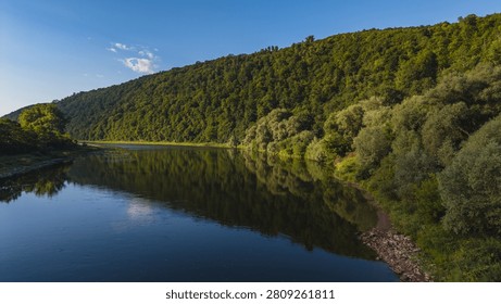 A river flows through a valley surrounded by hills and trees under a clear sky.