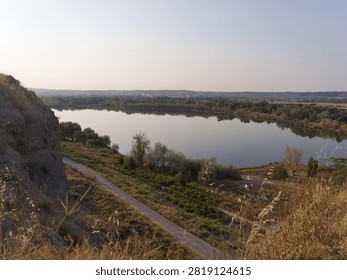 A river flows through a valley surrounded by green trees and hills. The sun shines down on the water, creating reflections. A path runs along the riverbank.