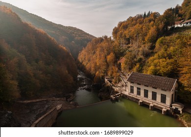 River With Green Colored Water, Runs Between Large Mountains Full Of Trees With Leaves Of Different Colors Due To Autumn. The Sun Illuminates The Hillside And An Old Hydroelectric Power Station.